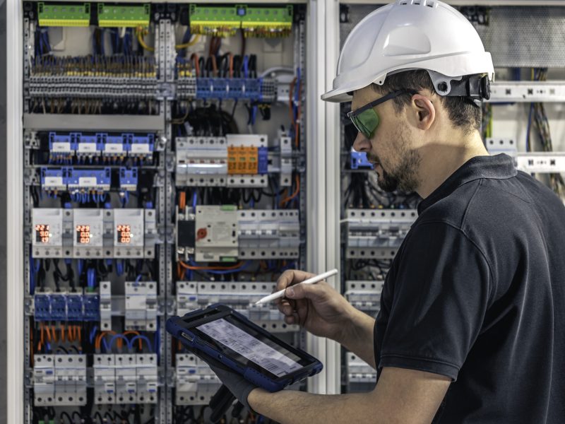 Man, an electrical technician working in a switchboard with fuses. Installation and connection of electrical equipment. Professional uses a tablet.